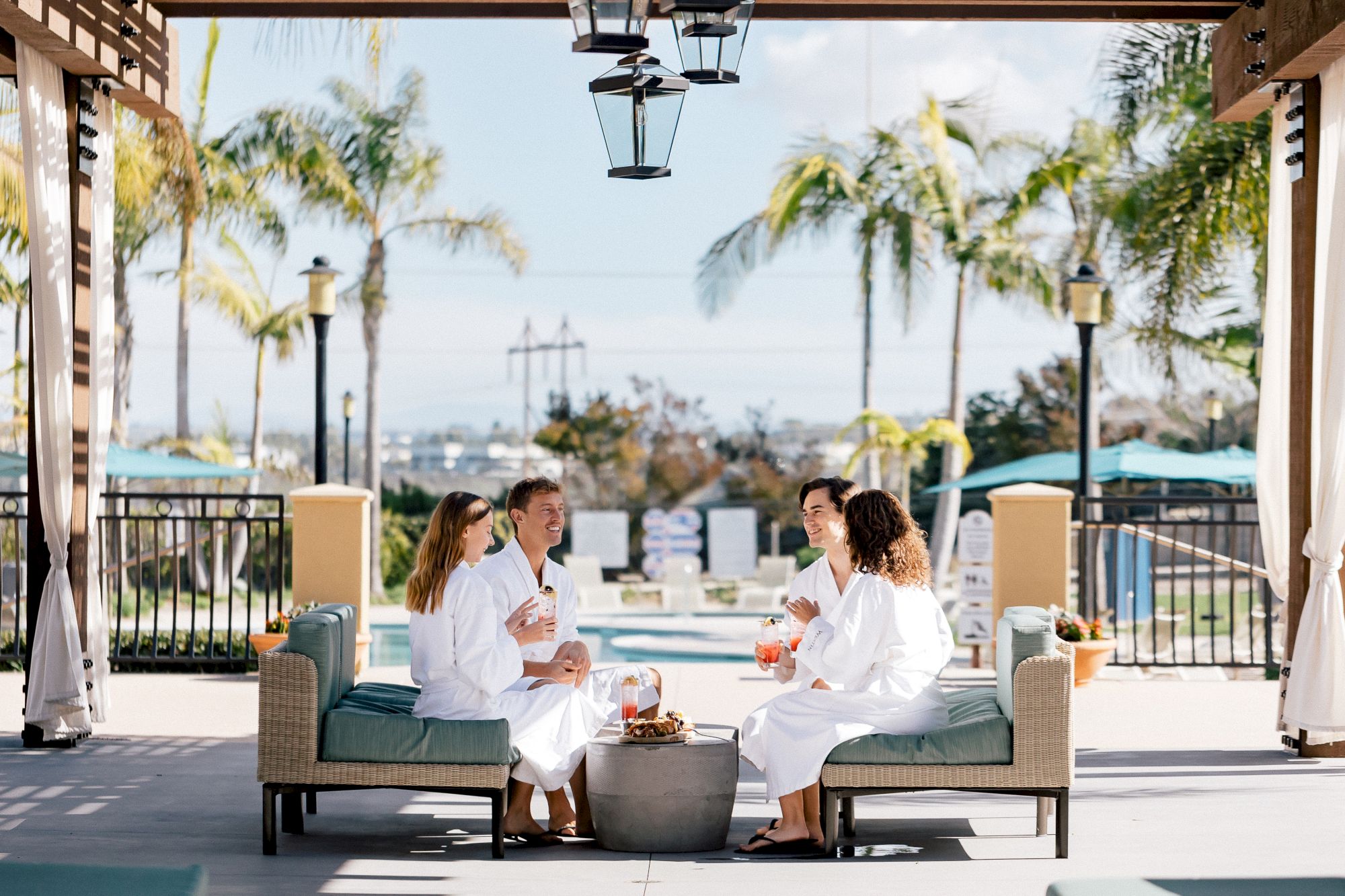 Four friends sit in white robes on a outdoor lounge, chatting and enjoying drinks under a shaded cabana with palm trees and a pool in the background.