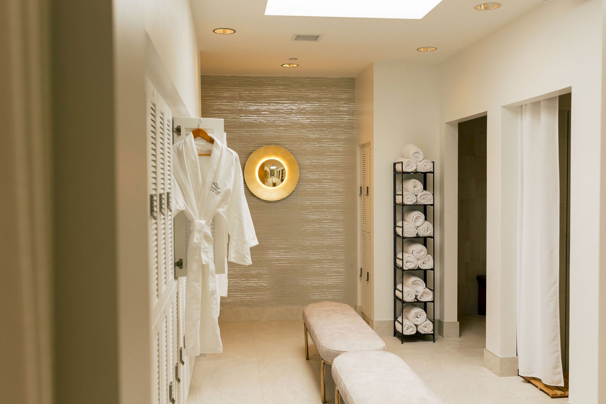The image shows a luxurious spa area with robes on hangers, a rack of rolled towels, soft lighting, a bench, and a round wall decoration.