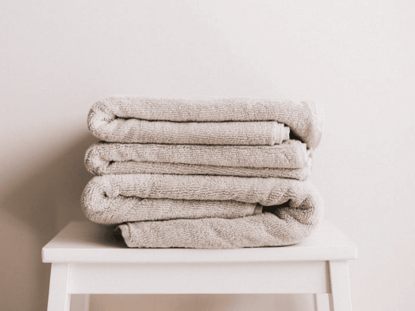 A stack of folded, beige towels is neatly placed on a white stool against a neutral wall.