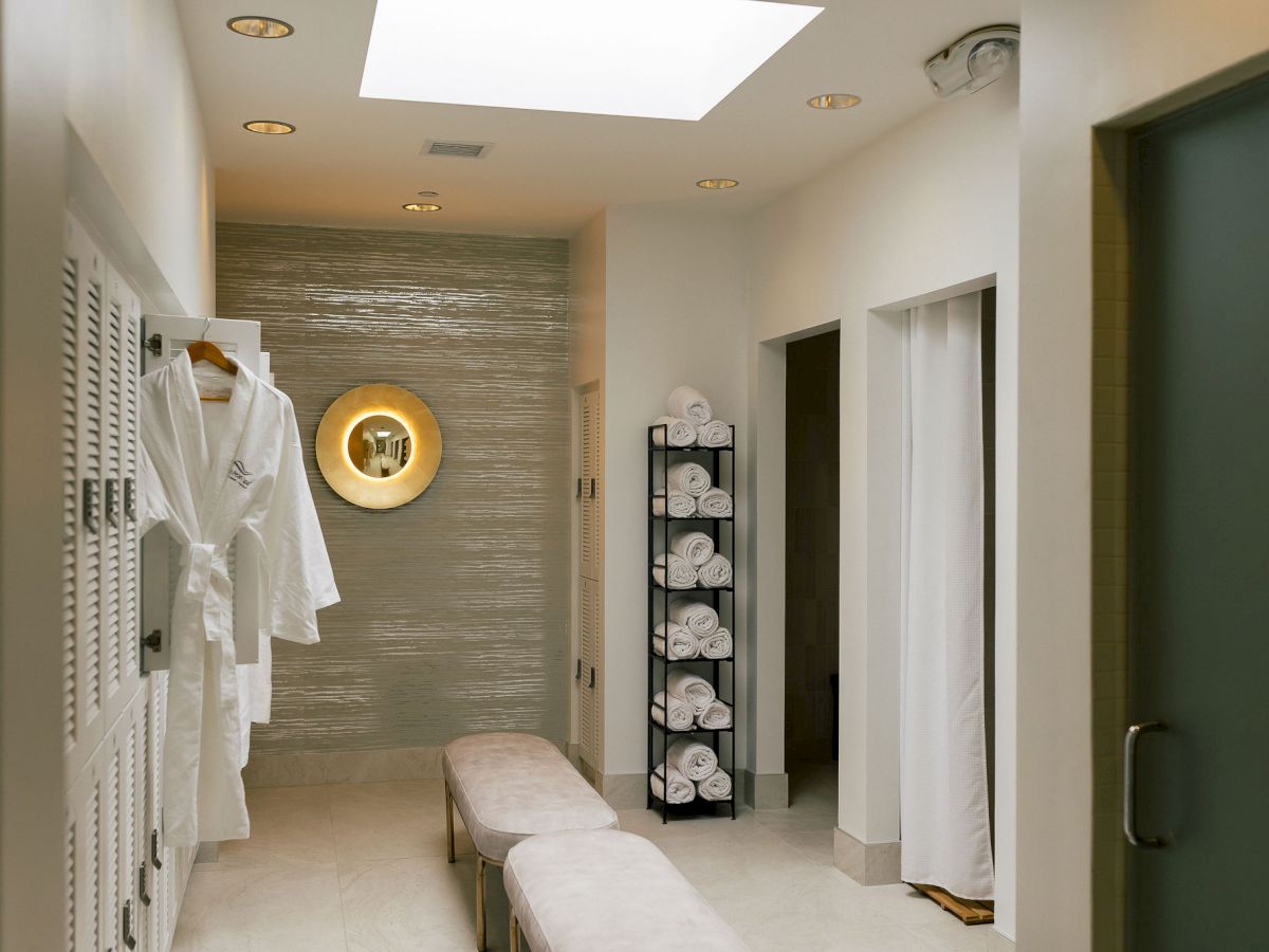 A spa dressing area with white robes on lockers, a cushioned bench, a circular mirror, and a shelf of folded towels in a calm, modern room.