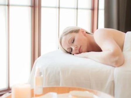 A person is lying on a massage table near a window, with a candle and some items on a small table in the foreground.