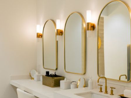 A modern bathroom with gold-framed mirrors, stylish lighting, a long white countertop, and a chair beneath the counter.