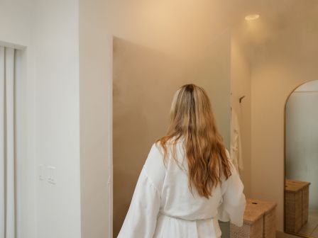 A person with long hair in a white robe is walking in a bright, minimalist room toward a doorway, with a curved mirror and wooden furniture visible.