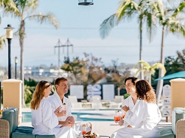 A group of four friends in white robes sits around a small fire pit on a resort patio, chatting and enjoying drinks under palm trees.